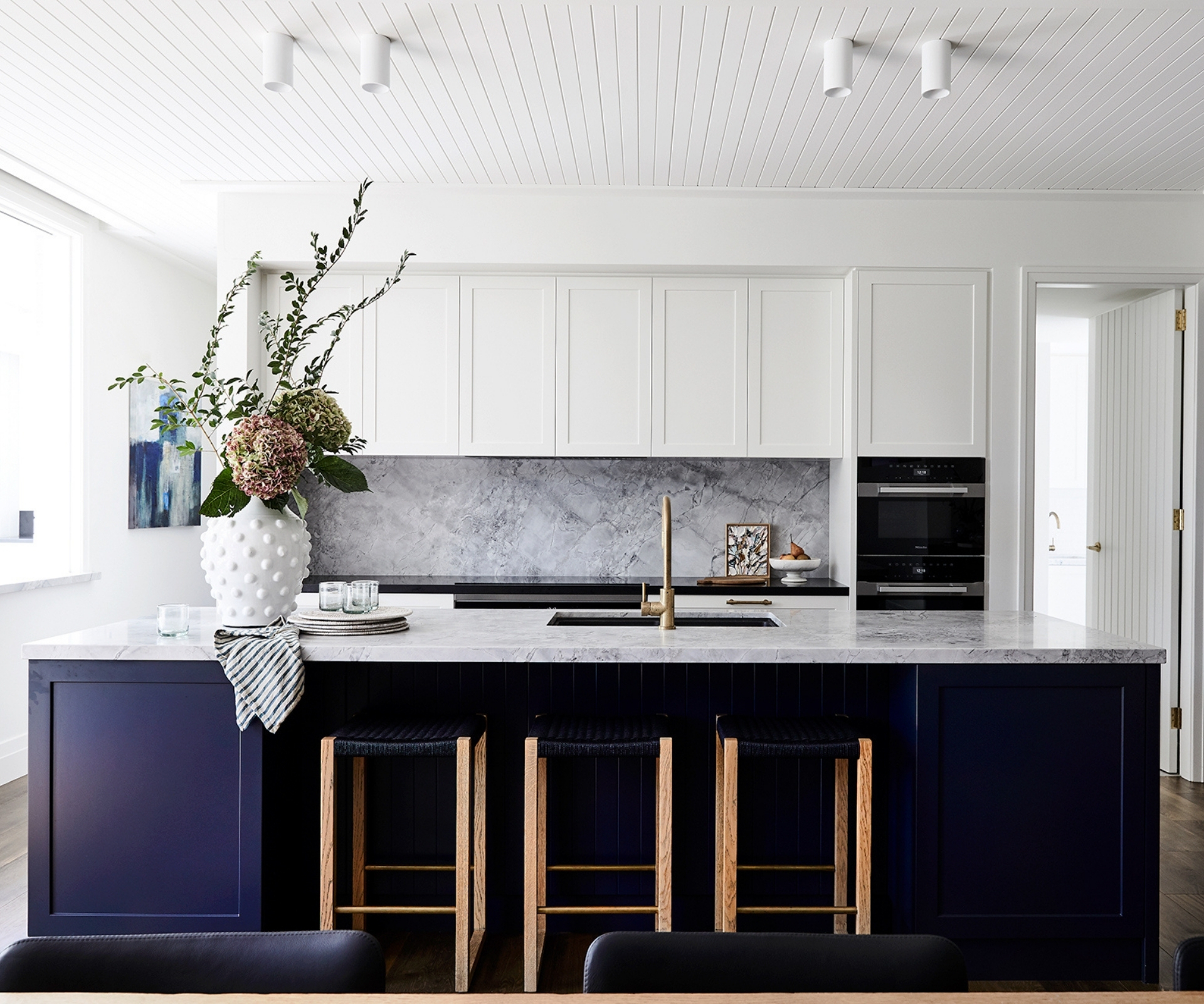 Modern kitchen with white cabinets, marble countertops, navy island, vase with flowers, and wooden bar stools.