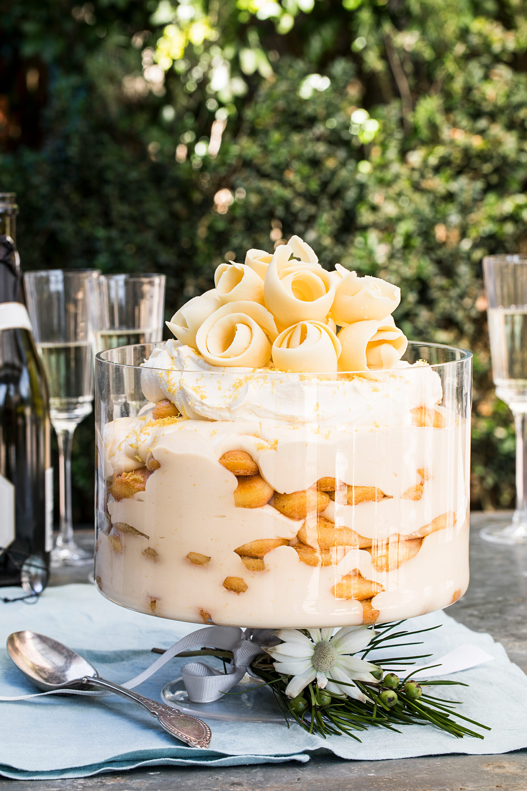 Limoncello trifle dessert topped with white chocolate curls in a clear bowl, surrounded by champagne glasses, on an outdoor table.