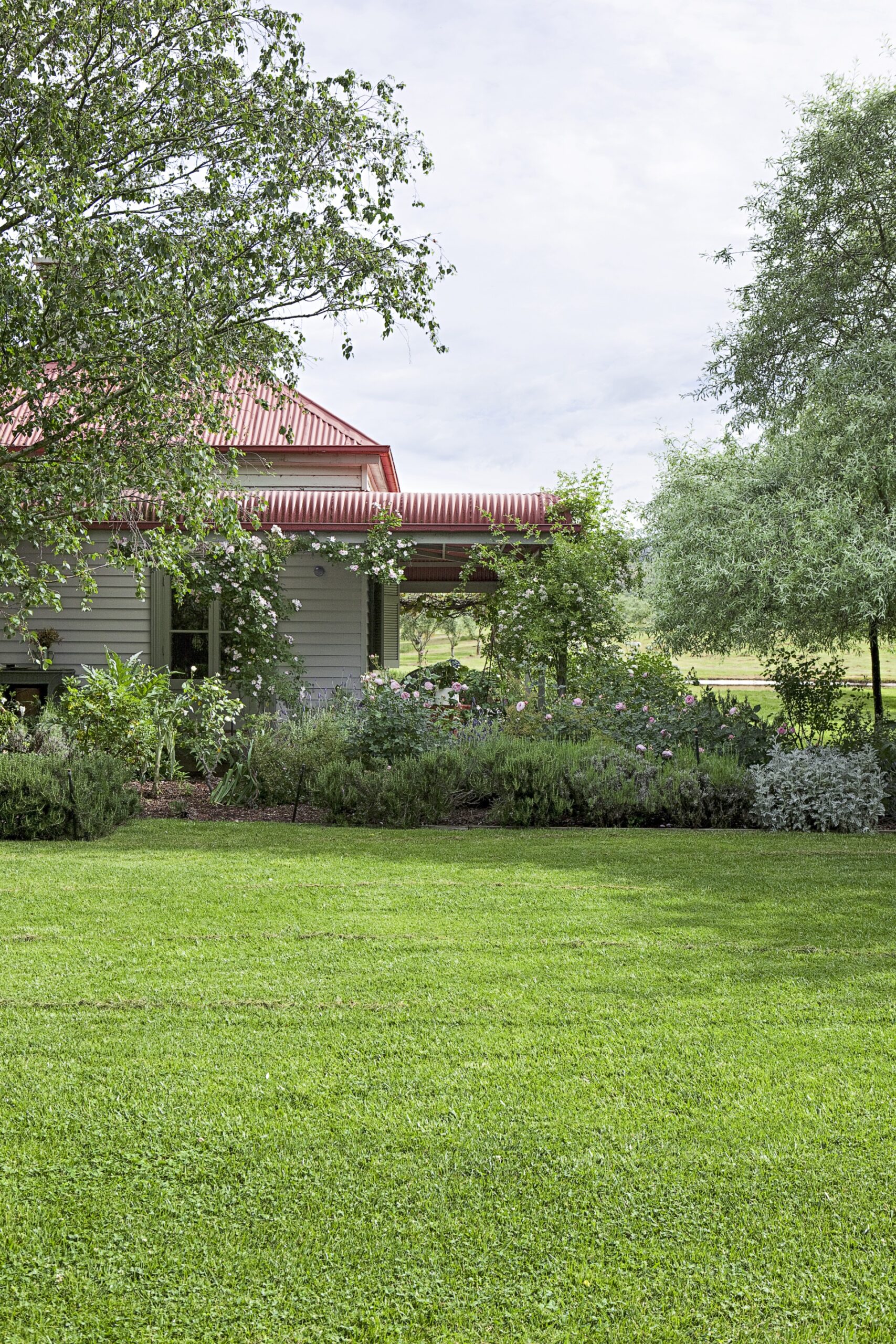 Red-roofed house surrounded by lush garden and trees, with a well-maintained green lawn in the foreground.