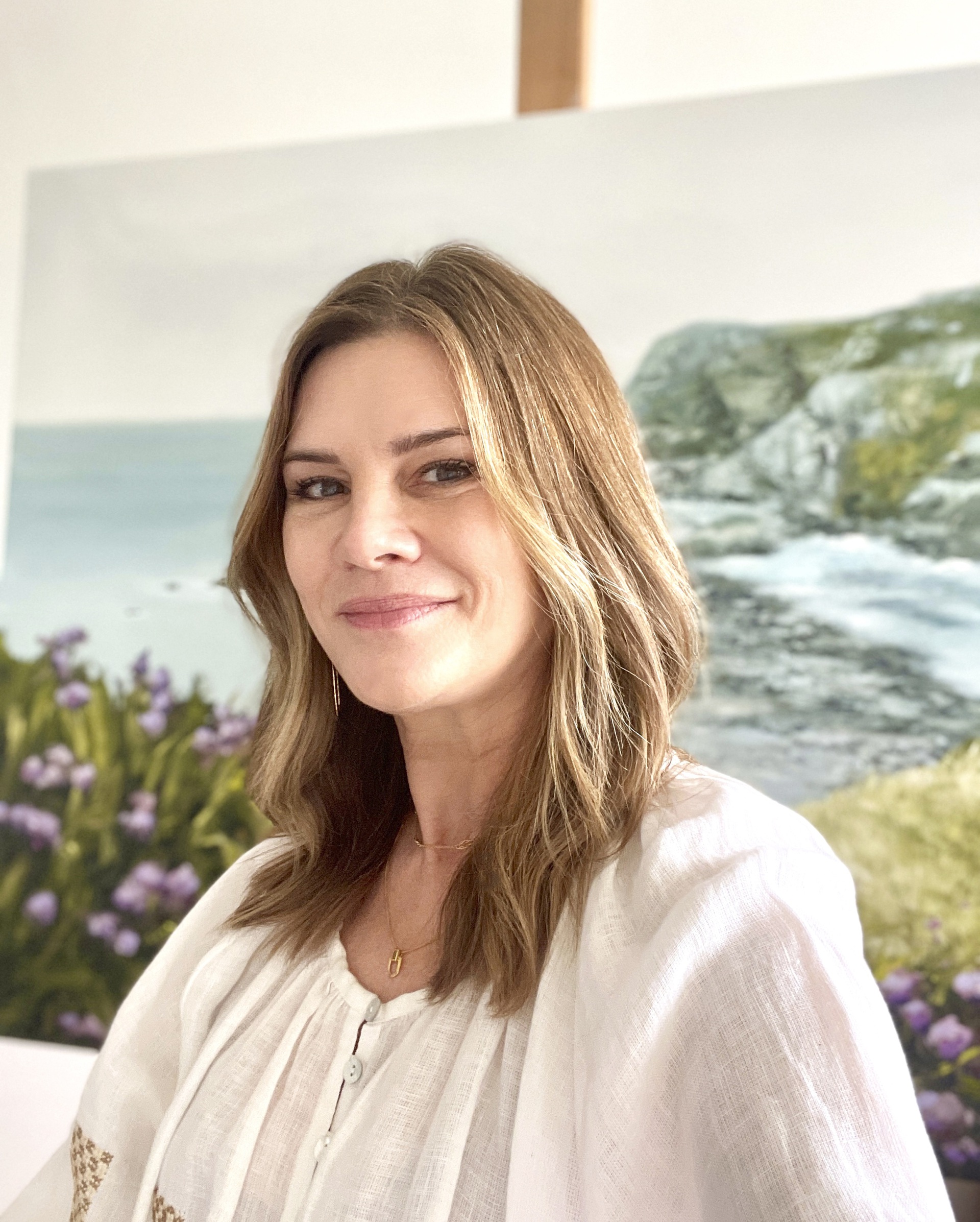 A portrait of artist Annie Hawkins by a coastal landscape painting.