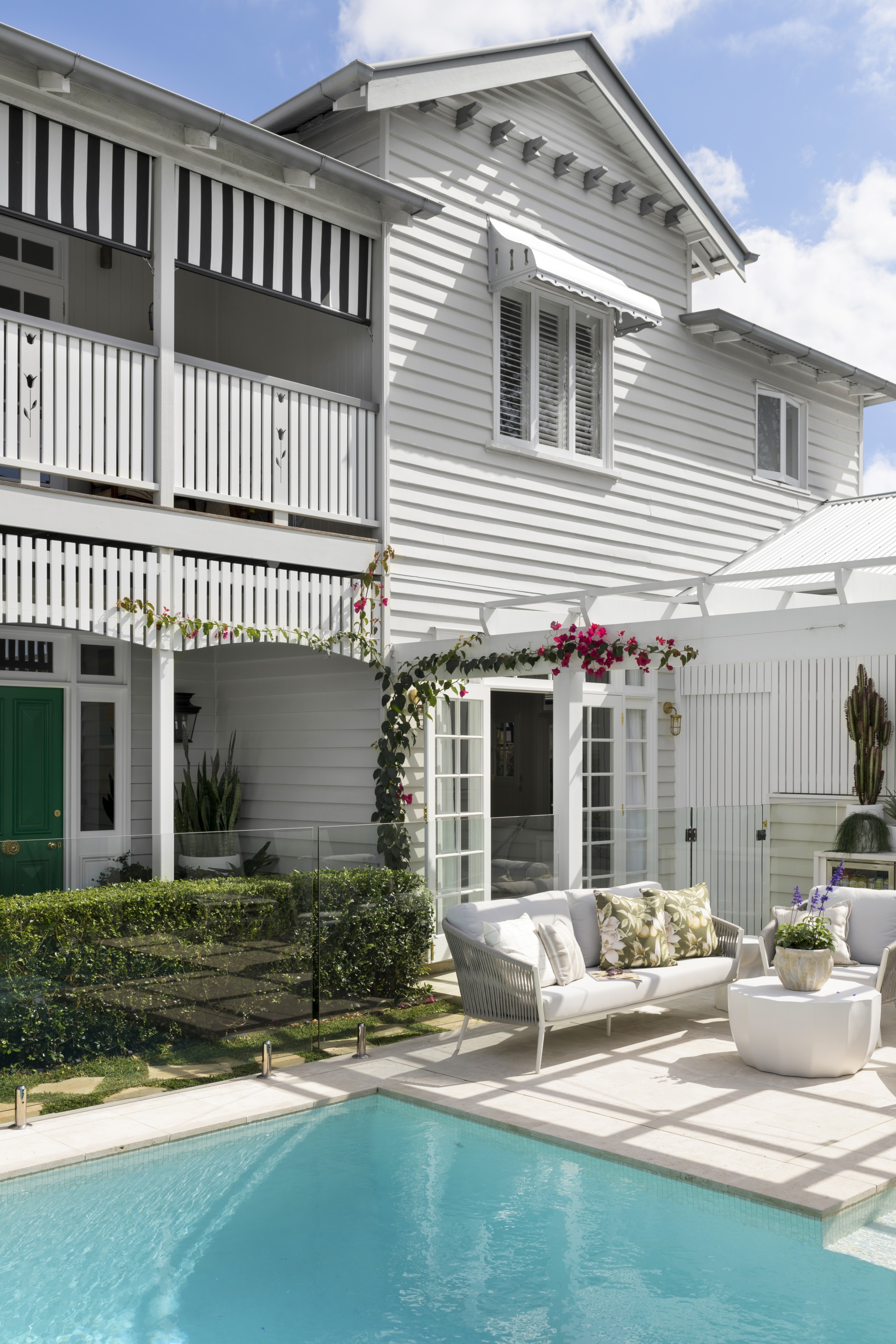 A white two-storey weatherboard cottage beside a pool and lounge chairs.