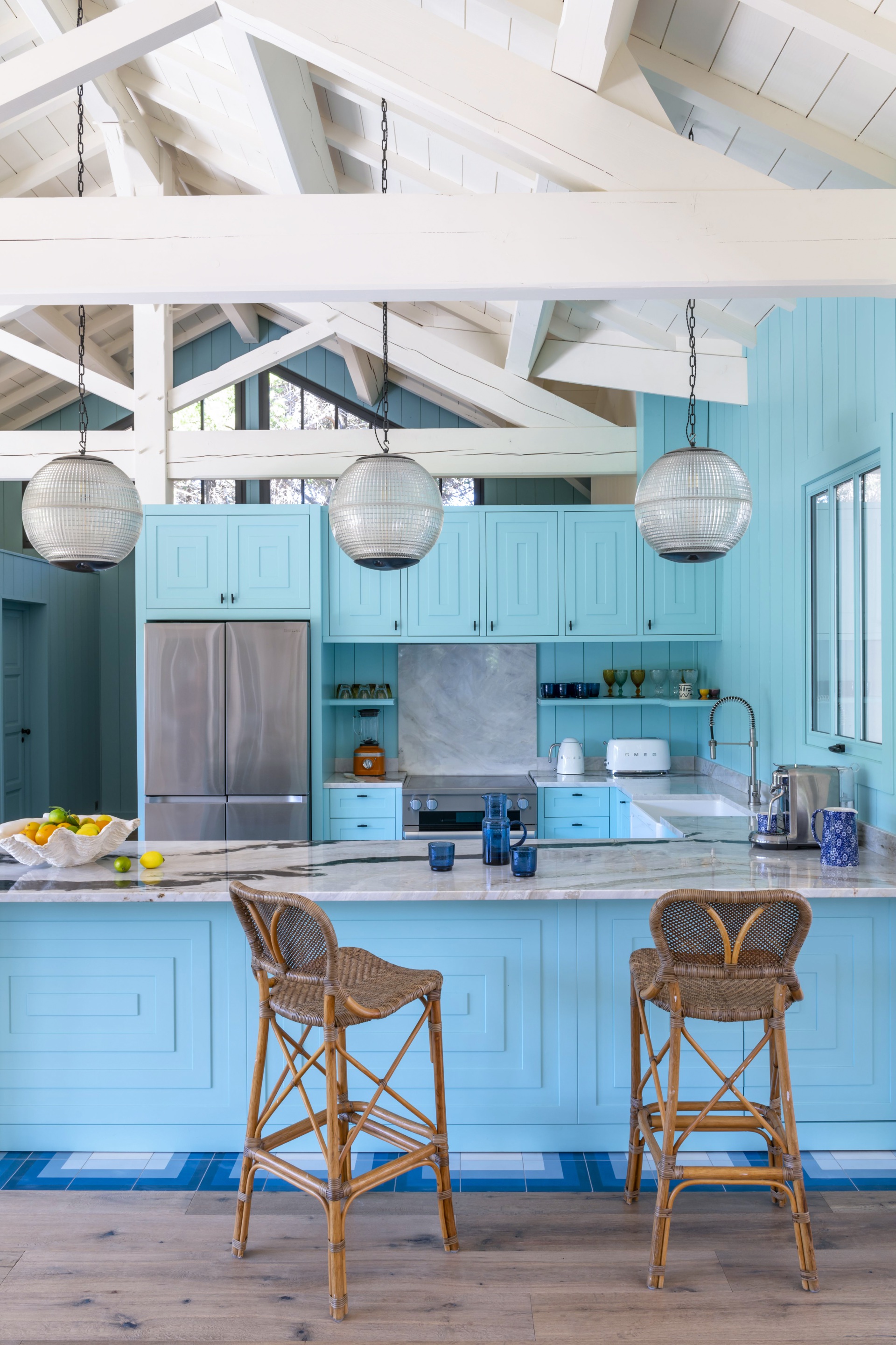 A coastal style kitchen with blue panelling, wicker stools and glass globe pendants.