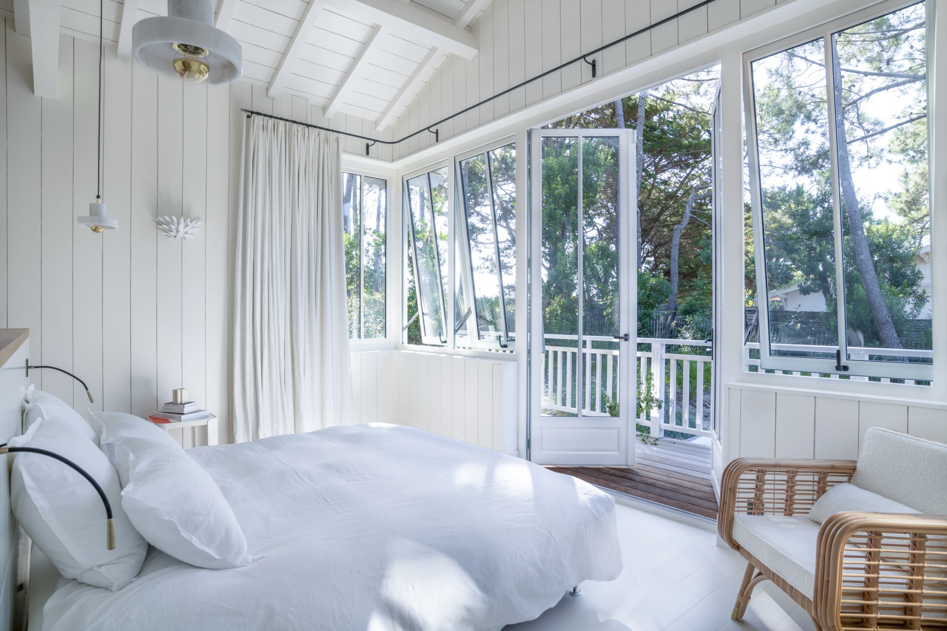 A white bedroom with a bed, wicker chair and balcony deck overlooking a garden.