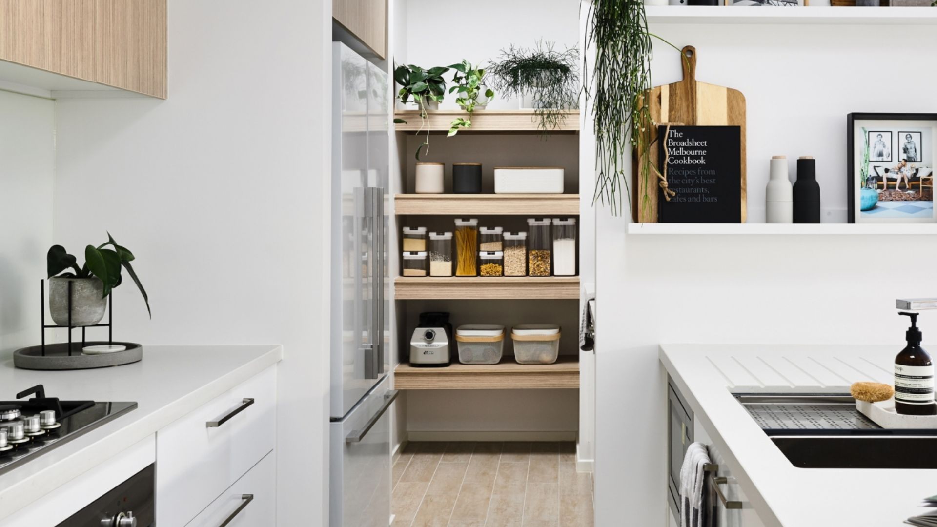 Modern kitchen with a pantry, plants, storage jars, and a cookbook. White countertops and wooden cabinets are visible.
