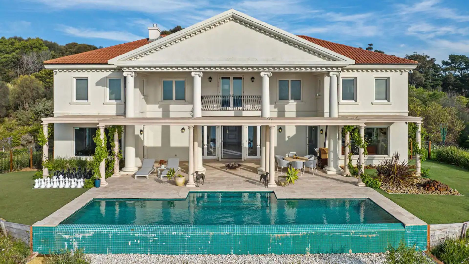 Grand white mansion with columns, red-tiled roof, patio, large swimming pool, and garden with giant chess set.
