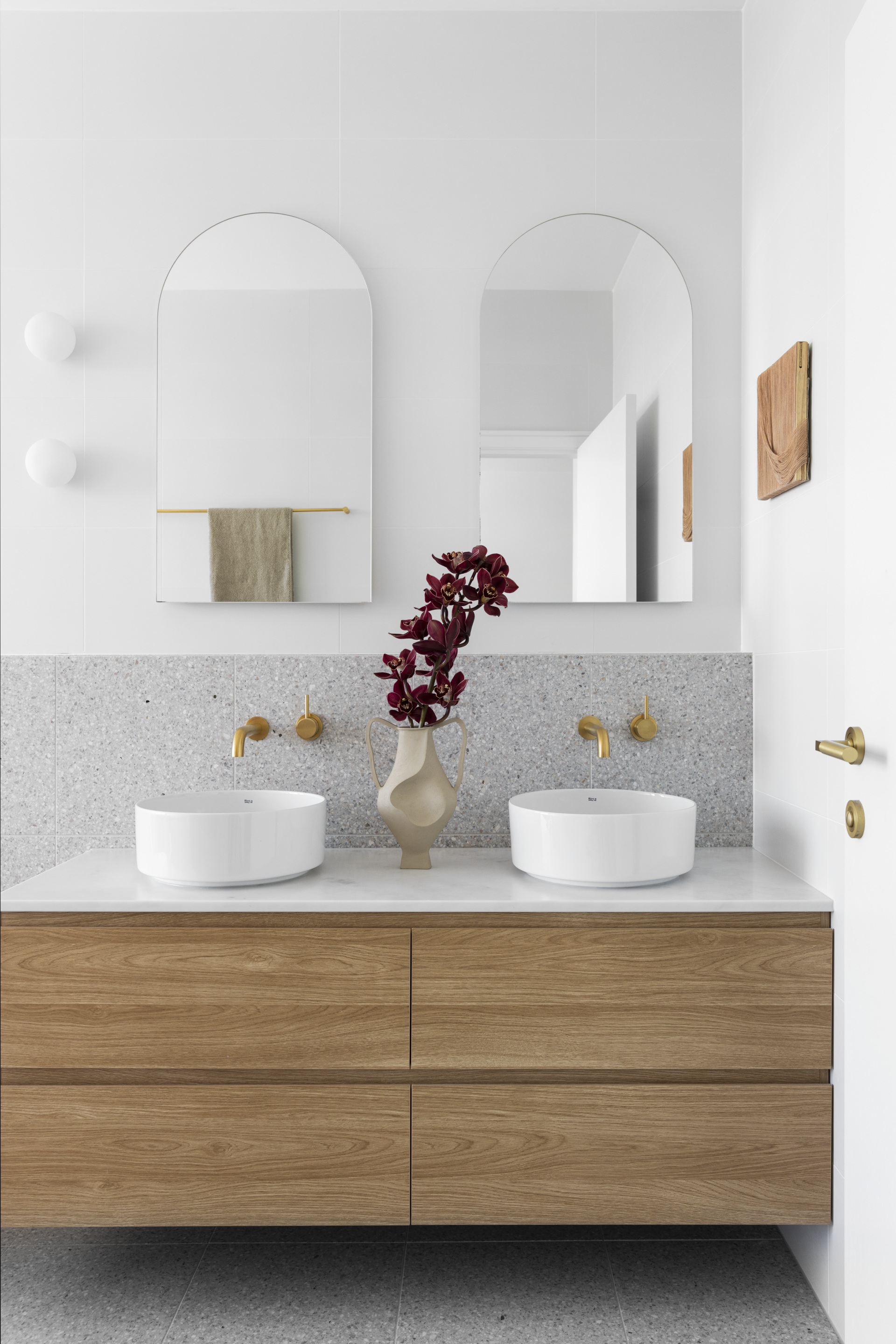 A white and grey ensuite with a timber vanity, gold tapware and arched mirrors.