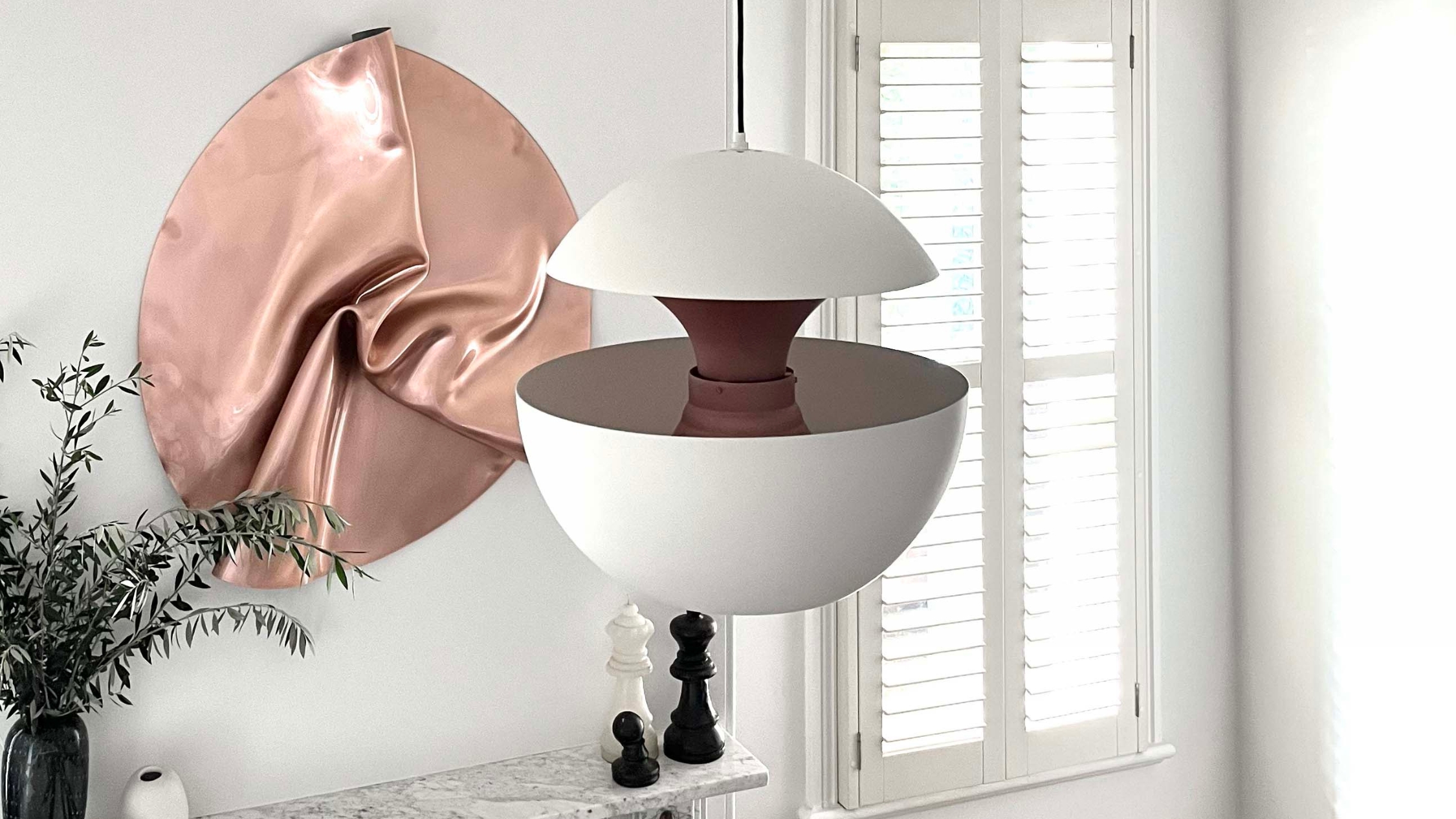 White modern pendant light, copper wall art, chess pieces, plant, and white shutters in a minimalist room.