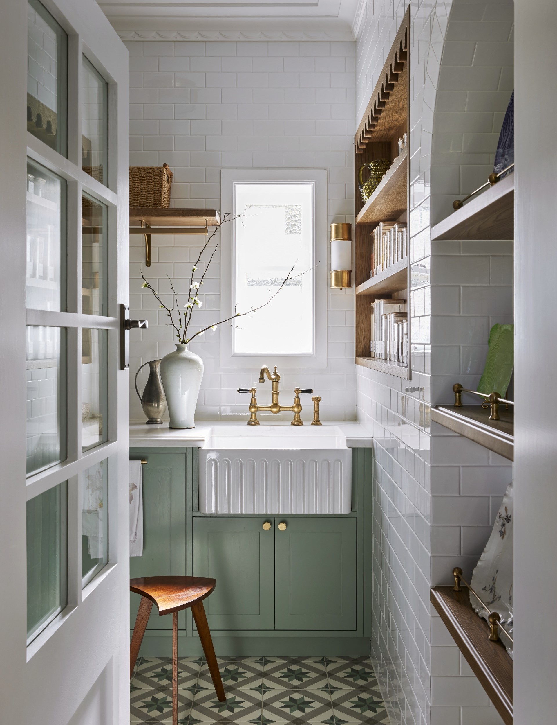 A butler's pantry with white subway tiled walls, a sizable fluted fireclay sink, hardware in Satin Brass and joinery finished in a mint green colour. Timber open shelving allows owners to display trays and dinnerware, plus containers filled with ingredients. The tiled flooring has a star pattern.